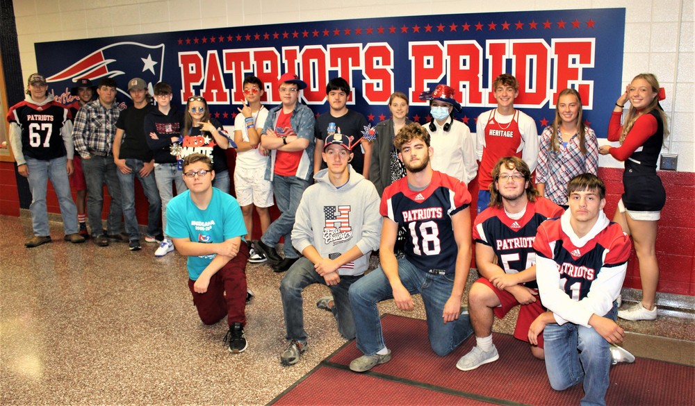 A large group of Heritage High School students pose in their red white and blue outfits in front of a large "Patriots Pride" sign that is on the wall.