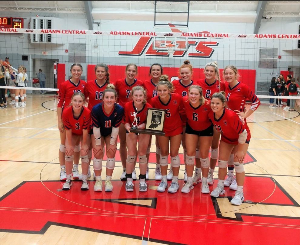 The 2022 Heritage girls volleyball team stands in front of the net on the Adams Central gym floor, after winning the ACAC tournament. 